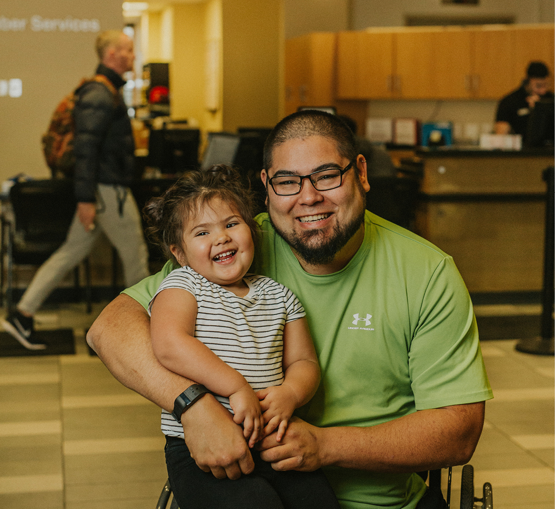 A man sits in a wheelchair with his daugther on his lap in the middle of a YMCA lobby.
