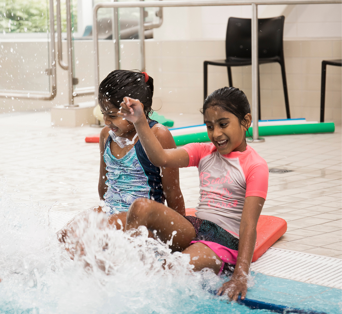 Two young girls wearing swim shirts and shorts sit at the edge of a pool while they kick their feet in the water.