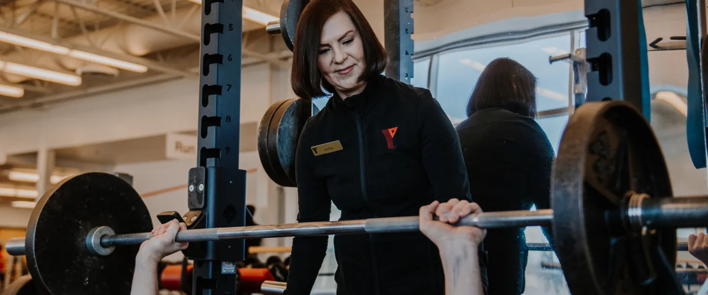 A female personal trainer stands behind a male member as he uses a barbell bench press.