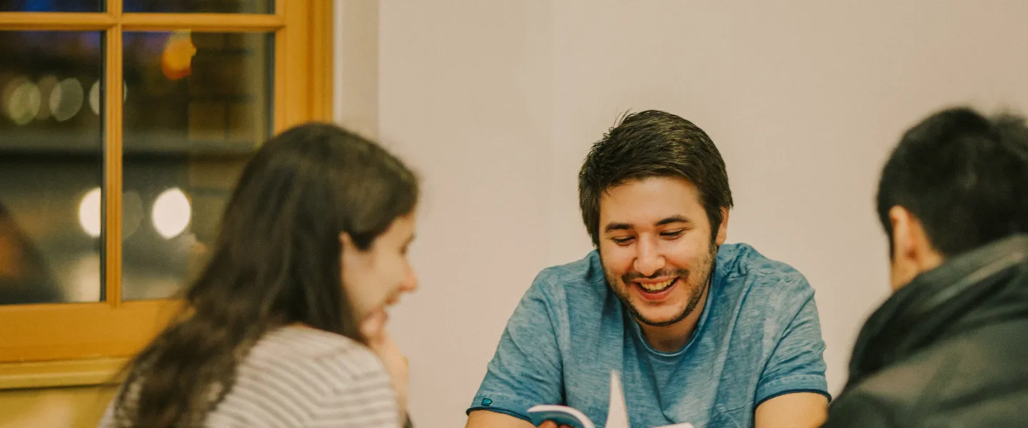 A teenage girl, teenage boy and male youth leader sit at a table reading a book together.