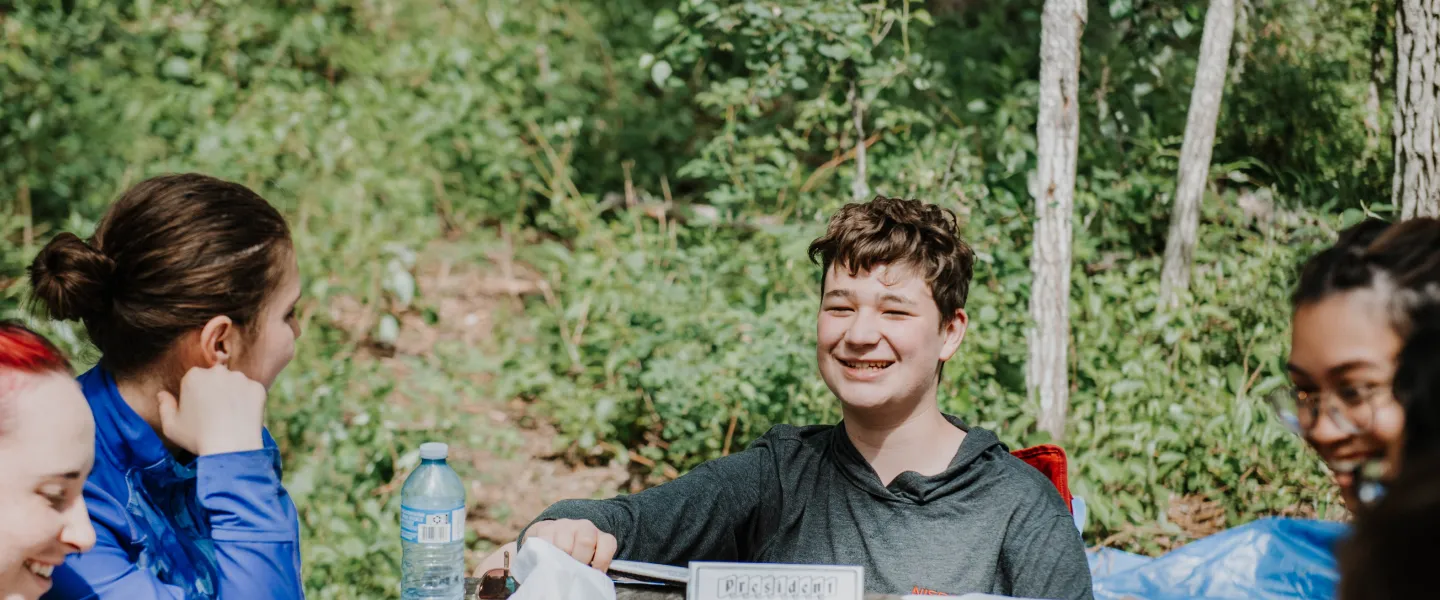 A group of teens play a card game on a wooden picknic table while camping.