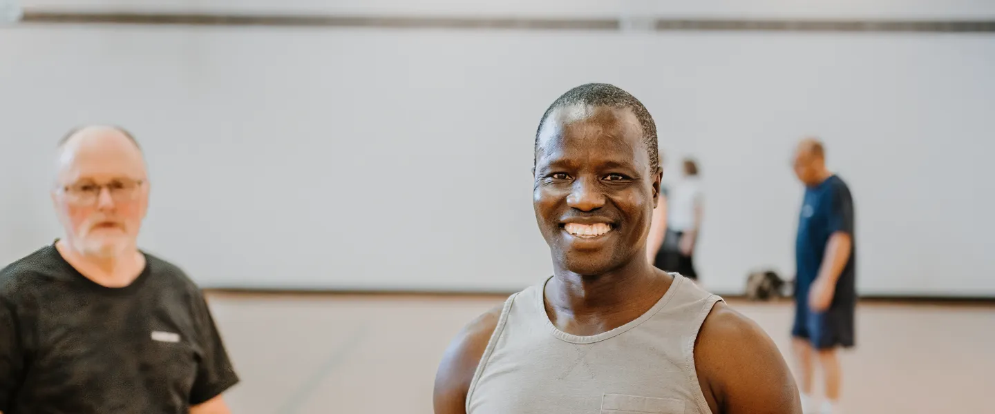 A smiling man stands in a gymnasium with members playing pickleball in the background.