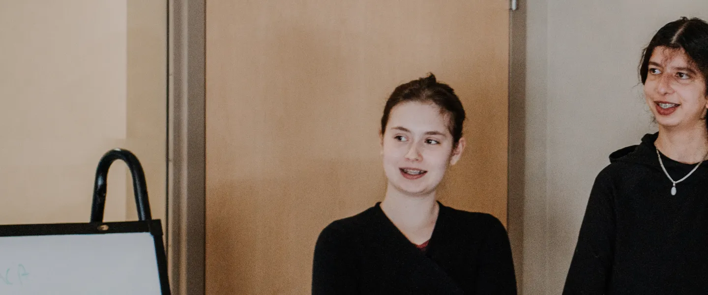 Two teenage girls stand infront of a classroom and listen to one of their peers talking.