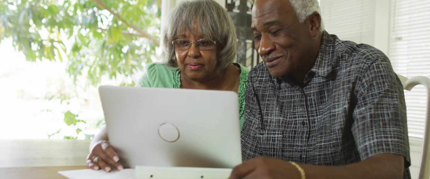 An older man and woman sit close together, using a computer at dinning room table.
