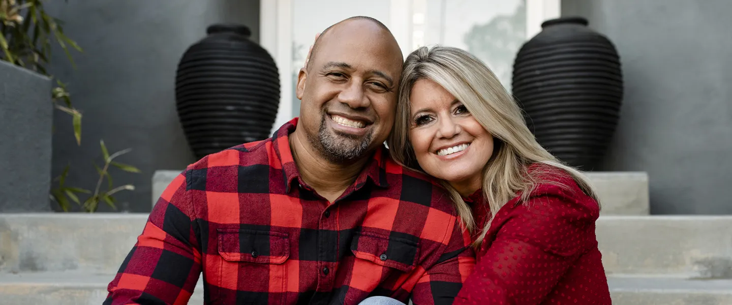 An interacial couple wearing red and black plaid shirts sit on the seps of their home.