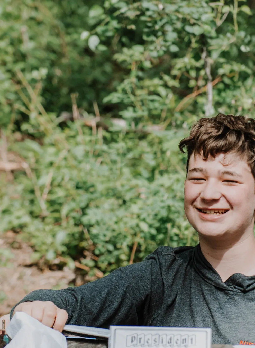 A group of teens play a card game on a wooden picknic table while camping.