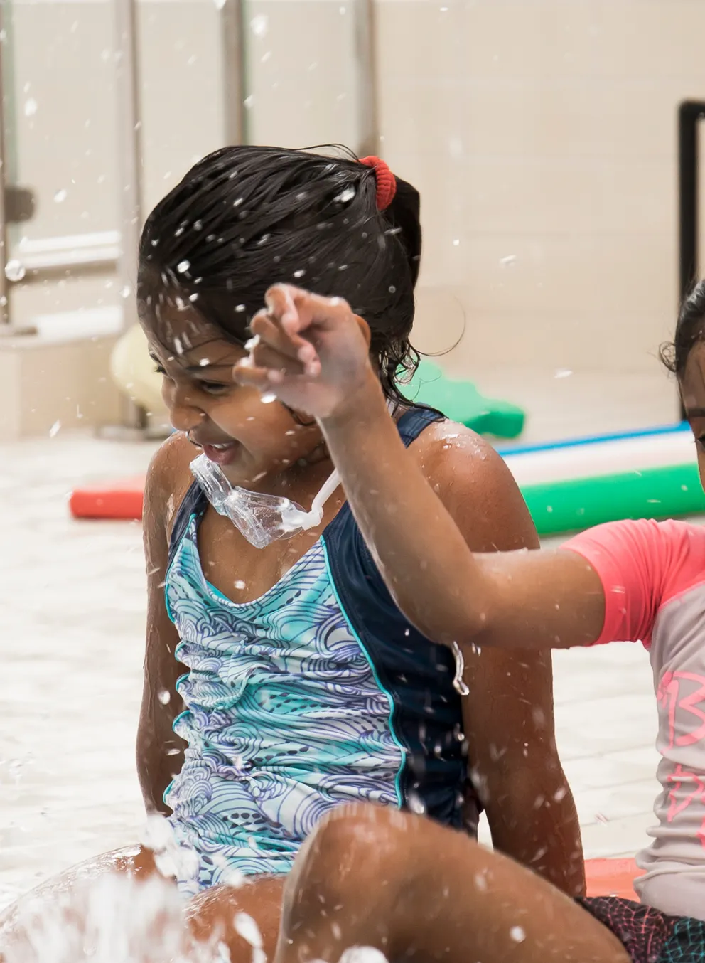 Two young girls wearing swim shirts and shorts sit at the edge of a pool while they kick their feet in the water.
