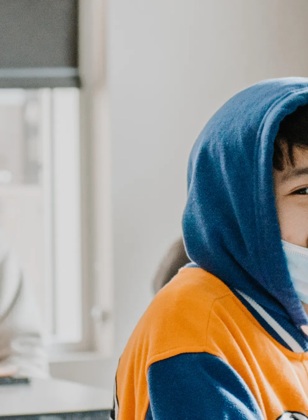 A teenage boy wears a hoodie with the hood up, sits at a table in a classroom with a laptop open.