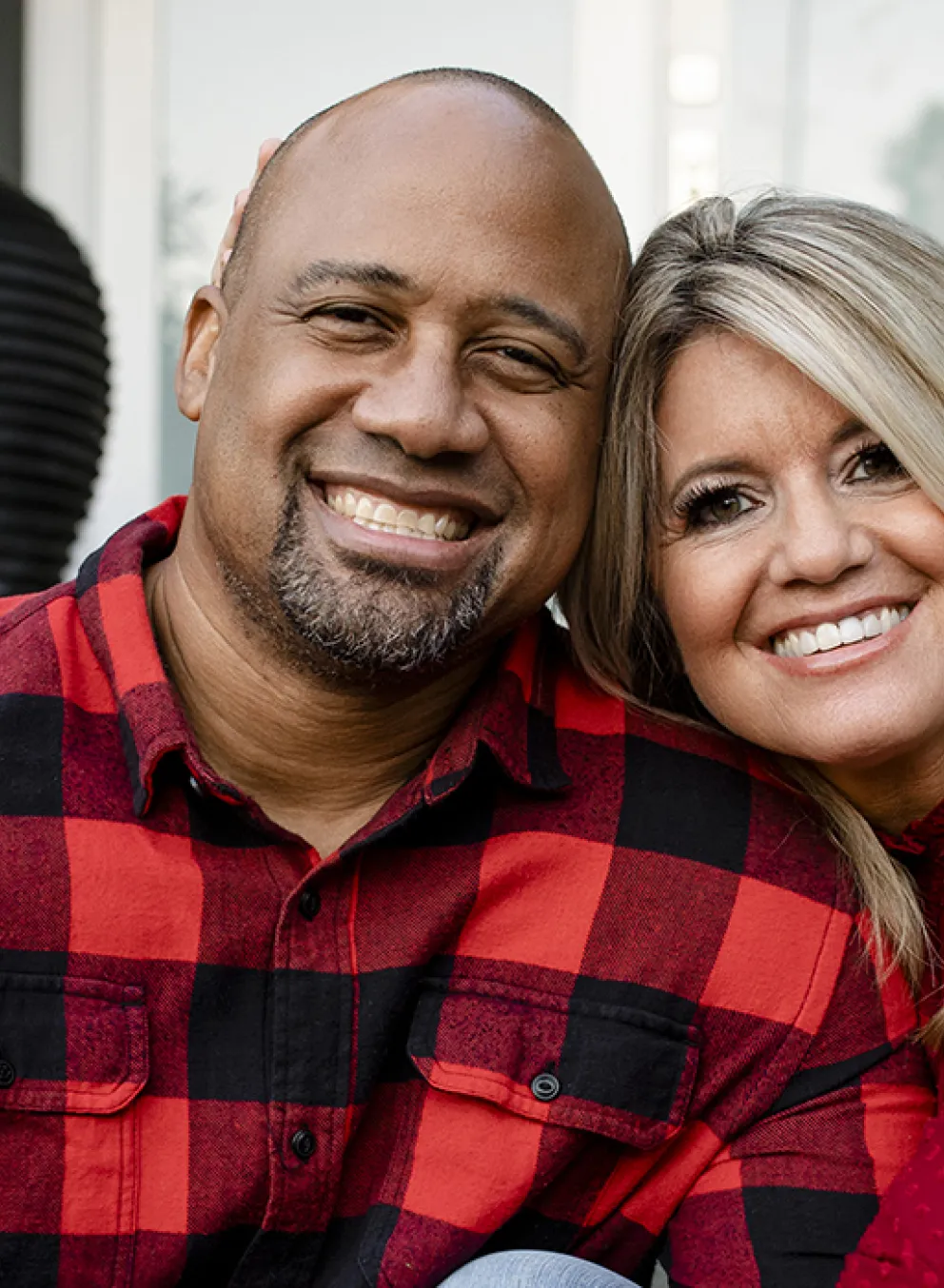 An interacial couple wearing red and black plaid shirts sit on the seps of their home.