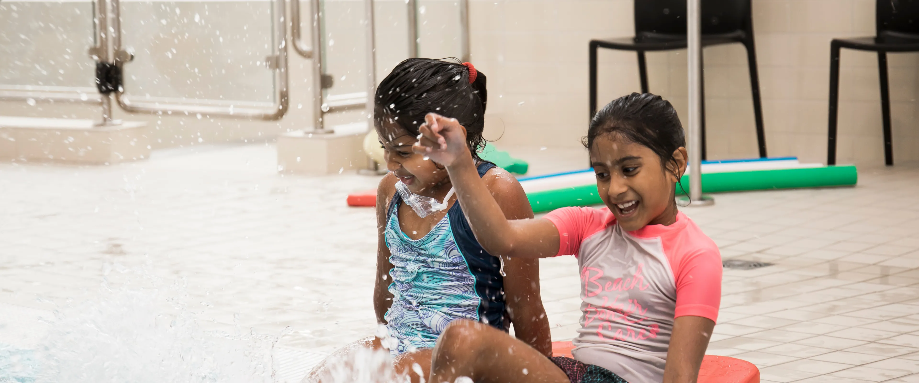 Two young girls wearing swim shirts and shorts sit at the edge of a pool while they kick their feet in the water.