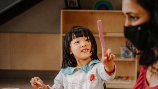 A young girl sticks a piece of felt to her finger and shows it to her early childhood educator.
