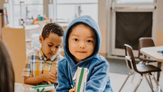 A young boy wearing his hood up shows off his craft that he made in a YMCA Child Care centre.