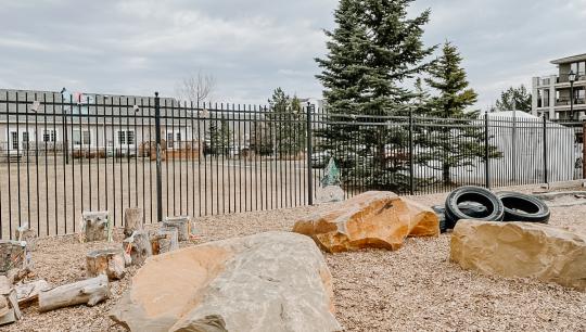 An outdoor fenced playspace with a tree, large rocks, tires and other playground style equiptment.