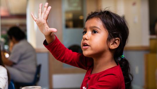 A young girl sitting on a small chair, in the middle of raising her hand and asking a question.