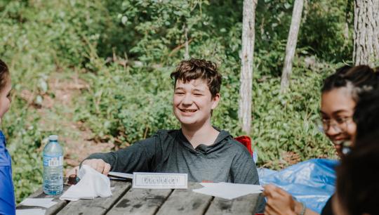 A group of teens play a card game on a wooden picknic table while camping.