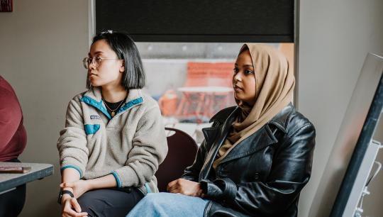 Two young women sit adjasent to each other in a brightly lit room while listening to a workshop.