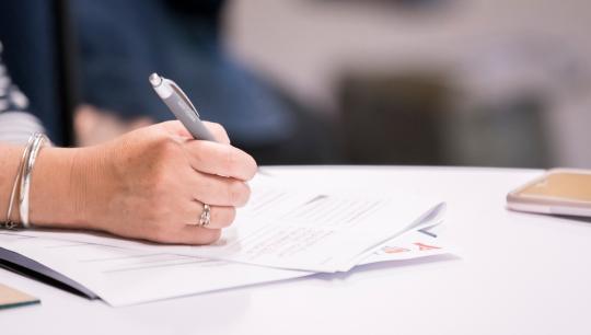 The hand of a woman using a pen to write notes on a small stack of papers during a workshop.