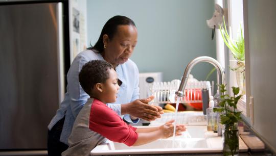 A mother and son wash their hands in a kitchen sink.
