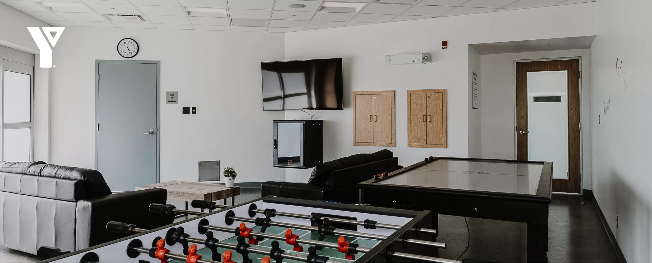 A room built for teens, featuring couches, a TV mounted on a wall with a gaming system, foosball table and air hockey table.