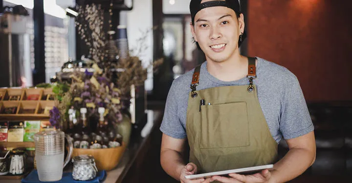 A young man wearing an apron holds a tablet while in a kitchen.
