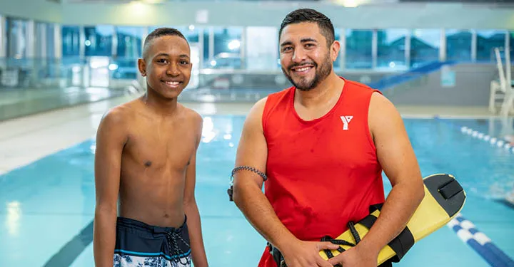 A teenaged boy and YMCA lifeguard stand together on a pool deck.